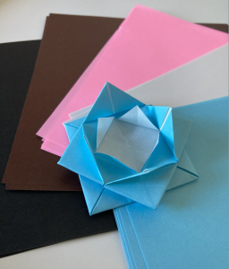 Light blue origami lotus flower on stack of origami paper in trans and progress pride flag colors