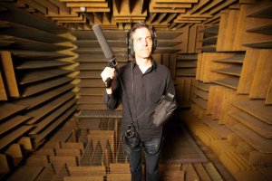 Sam Green, wearing headphones and carrying a microphone, in front of a background of wooden sound baffles
