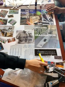 The hands of two seated figures work at a table full of cutout articles about Detroit