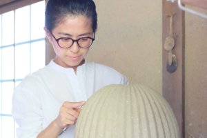 Yoshiko Takahashi, a woman with dark hair wearing a white shirt and black-framed glasses, carves a clay sculpture with a tool.