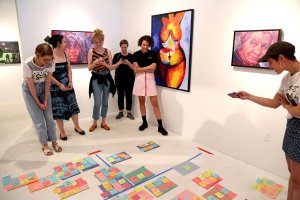 A group of women stand in a gallery, interacting with a grid of post-it notes arranged on the floor