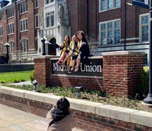 Three women sit on the Michigan Union sign in graduation gowns, tossing their mortar boards at a photographer crouched on the sidewalk.