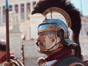 A male Roman soldier reenactor wearing a plumed helmet and armor and holding a weapon.