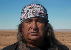 A man wearing a multicolored, patterned bandana over his long, grey hair in front of a desert landscape, photographed from the chest up.