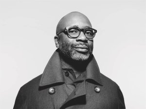 Theaster Gates, a Black man with a shaved head and salt and pepper beard, wearing black-rimmed glasses and a pea coat, looks at the camera in a 3/4 profile shot.
