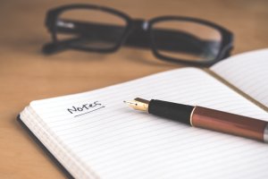 a pen on an opened notepad. the word notes is written on one of the pages and there's black glasses next to the notepad.