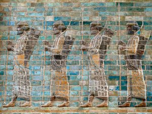 Blue, black, and orange brick relief image of four bearded soldiers in elaborately decorated clothing walking with long spears, a bow, and a quiver.