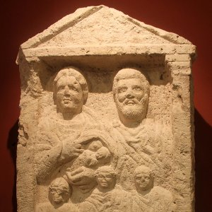 Tan carved stone grave marker showing a man and a woman breastfeeding an infant, with three more children in front of them. Tan carved stone grave marker showing a man and a woman breastfeeding an infant, with three more children in front of them.