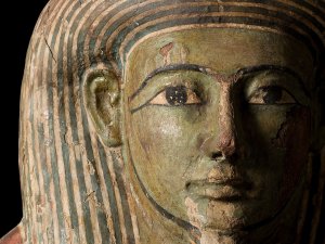 The head of a detailed, wooden coffin. Its face is green in color, while blueish stripes line the headpiece.