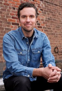 John Hendrickson has brown hair and brown eyes. He's wearing a denim button-up shirt and sitting in front of a brick wall.