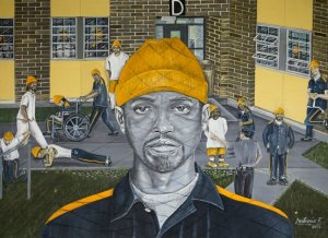 In this drawing, a man wearing an orange hat and jumpsuit is pictured in the foreground, with the shadows of wire mesh across his face. Other inmates exercise and walk through a courtyard behind him.