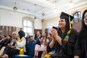 Student sitting and wearing cap and gown clapping at graduation ceremony.