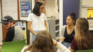 Four people in a circle are talking. One person is off to the side not in the circle. One person is standing. All people are looking at the person who is talking.