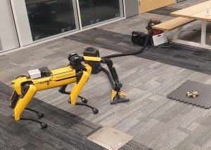 A yellow quadruped robot with a grasping arm reaches for the end of a vacuum hose to drag it over an obstacle and toward a mess of blocks.