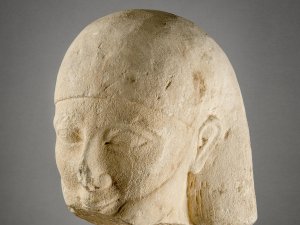 Limestone sculpture of a male head with an Egyptian hairstyle. Much of the figure’s chin is broken off.