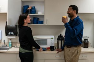 A woman puts away dishes while speaking to a man eating an apple.