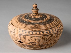 Pyxis with a lid made from buff clay and slip with brown-black glaze. The lid is decorated with concentric circles, while the box has varied patterns of dots and lines.