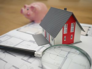 Small red and white model house with grey roof pictured with a pink piggy bank, a magnifying glass, and blueprints