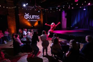 Photo during the UMS Fall 2023 Residency at the Ypsilanti Freighthouse, a Drag Queen can be seen looking fabulous in pink on stage with arms extended and the audience going wild.