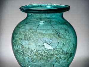 Blue-green glass urn with a piece missing and some cracks on the surface.