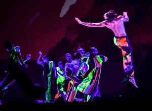 Seven dancers from Cloud Gate Dance Theatre company are seen wearing bright neon colors dancing on a dimly lit stage. They are glowing from UV lighting.