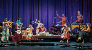 Members of the Silkroad ensemble performing on stage in front of a purple backdrop. A variety of instruments can be seen including Rhiannon Giddens is seater to the left of the stage playing banjo.