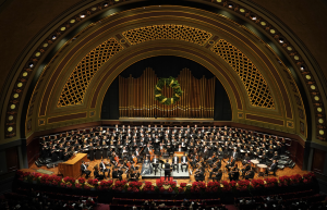 UMS's presentation of Handel's Messiah in Hill Auditorium 2023. The stage is full with the UMS Choral Union upstage on risers, the Ann Arbor Symphony orchestra upstage, Scott Hanoian can be seen conducting. The stage is decorated with Poinsettias a long the front edge, and a giant holiday wreath hangs above everyone.