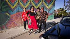 Four members of La Santa Cecilia pictured wearing bright colors, posing by a car in an alley with an abstract mural behind them.