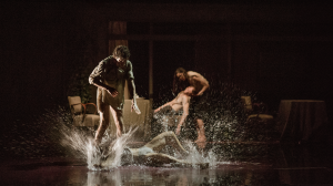 Image from mid performance, dancers can be seen on a very dimly lit stage splashing around in water.