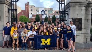 A large group of University of Michigan students pose with a blue Michigan flag, bearing a maize "Block M" in front of the Trustees Gate