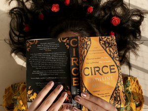 A woman lies on the ground reading “Circe” by Madeleine Miller. Her hair is splayed around her head and decorated with flowers.