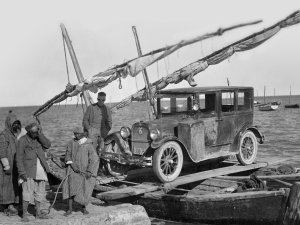 A 1920s sedan loaded atop a makeshift ferry consisting of fishing boats connected by ropes and planks. To the left stands a worker from the University of Michigan expedition and three Tunisian men.