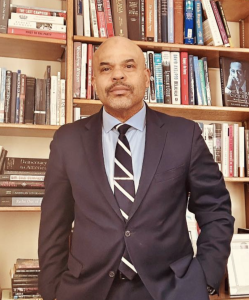 Photograph of the author Saladin Ambar, a Black man wearing a blue suit and tie against a background of bookshelves