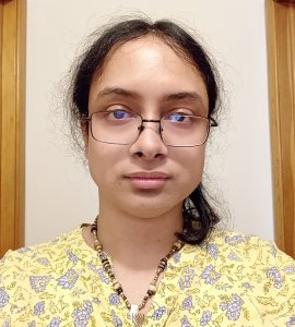 Close up headshot of Aniruddha Dutta wearing a yellow shirt, beaded necklace, and dark hair pulled back