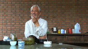 Smiling individual with white hair and a white beard standing over a counter with various kitchen dishes scattered around
