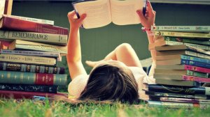 Woman lying on her back in grass, reading a book, with stacks of books on either side of her.