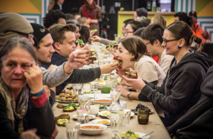 Several people sit on either side of a long, narrow table. Diners feed each other food from across the table and engage in conversation. Image from a previous collaboration between artists Dawn Weleski and Jen Rae.