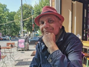 Man wears red cap and blue jacket while sitting at bustling sidewalk cafe' in Berlin, Germany.