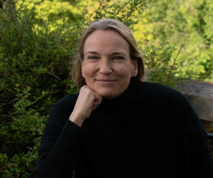 Grinning woman with light brown hair and a black turtleneck rests her face on her fist surrounded by greenery.