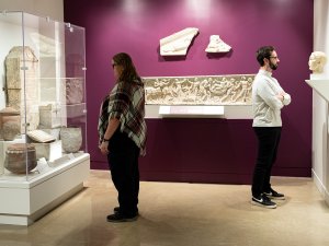 Two people stand in the Kelsey Museum’s Roman galleries looking at objects relating to funerary culture, including ash urns. In the background is a long, marble sarcophagus.