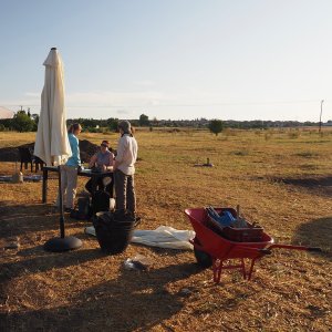 Members of the team prepare for work in the morning at Pella, Greece: a group of students gathers around a table, with supplies, including buckets, an umbrella, and a wheelbarrow, visible in the foreground.