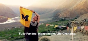 A young woman wearing a scarf stands in front of scenic mountains holding a U of M flag