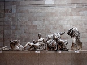 View of the Elgin Marbles at the British Museum, showing a row of white marble sculptures: the head of a horse, a nude reclining man with his hands and feet broken off, two headless figures in drapery seated together, and a headless, armless statue of a standing woman.