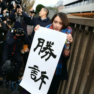 A woman wearing a colorful scarf holds a sign in Japanese while standing next to a group of photographers