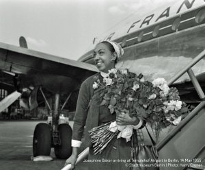 Young woman, in 1955 Berlin, stands in front of air france airplane holding a large flower bouquet.
