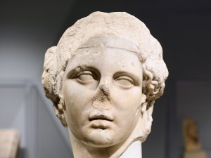 White marble head of Bacchus/Dionysus, whose curly hair is pulled back in a low bun and whose nose is broken off. White marble head of Bacchus/Dionysus, whose curly hair is pulled back in a low bun and whose nose is broken off.
