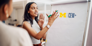 A presenter gestures towards a flip chart displaying the CEW+ logo while engaging an audience in a discussion. A presenter gestures towards a flip chart displaying the CEW+ logo while engaging an audience in a discussion.
