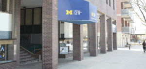 The entrance of a brick building featuring a blue awning with the text 'M | CEW+' visible. A sign and staircase are located to the left of the entrance, and a person is walking on the sidewalk.