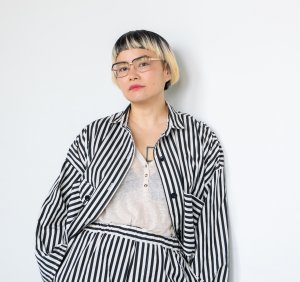 Headshot of Anne Ishii. She is leaning back against a white wall and wearing a black and white striped shirt and pants. She has short blond hair and glasses.