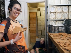 Students working on the Kea project in a storage building. Lauren Alberti holds up a triangular object for the camera, while a group of additional objects are scattered on a table in front of her.